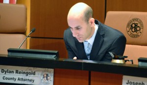 County Attorney Dylan Reingold at the dais during a County Commission meeting. The logo of County government is positioned on his nameplate and embroidered onto his seat back. 